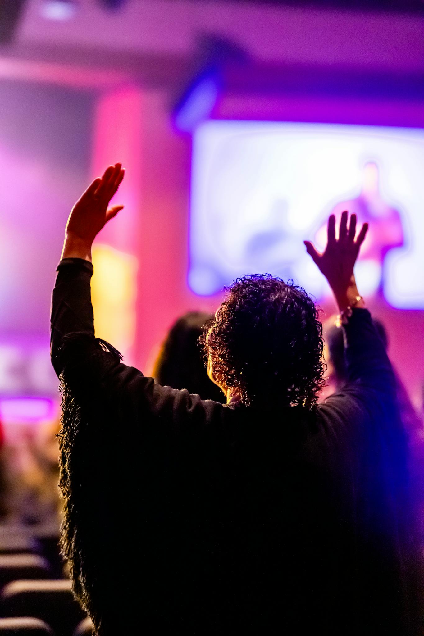 A person raising their hands in worship, silhouetted against colorful stage lights in a church setting.